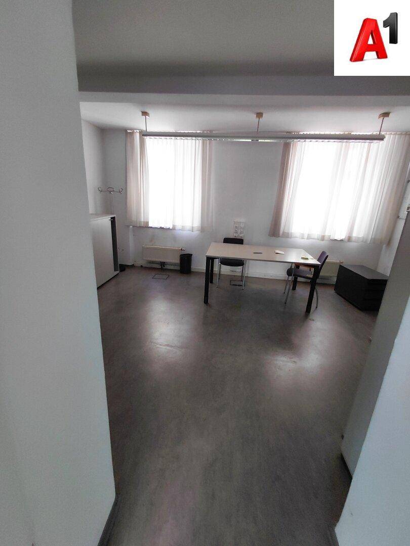 Kleineres Büro oder Pausenraum mit Tisch, Stühlen, Kühlschrank und Fenstern mit Vorhängen.
