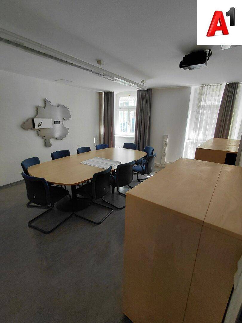 Besprechungsraum mit großem ovalen Holztisch, blauen Stühlen und Fenstern mit Vorhängen.