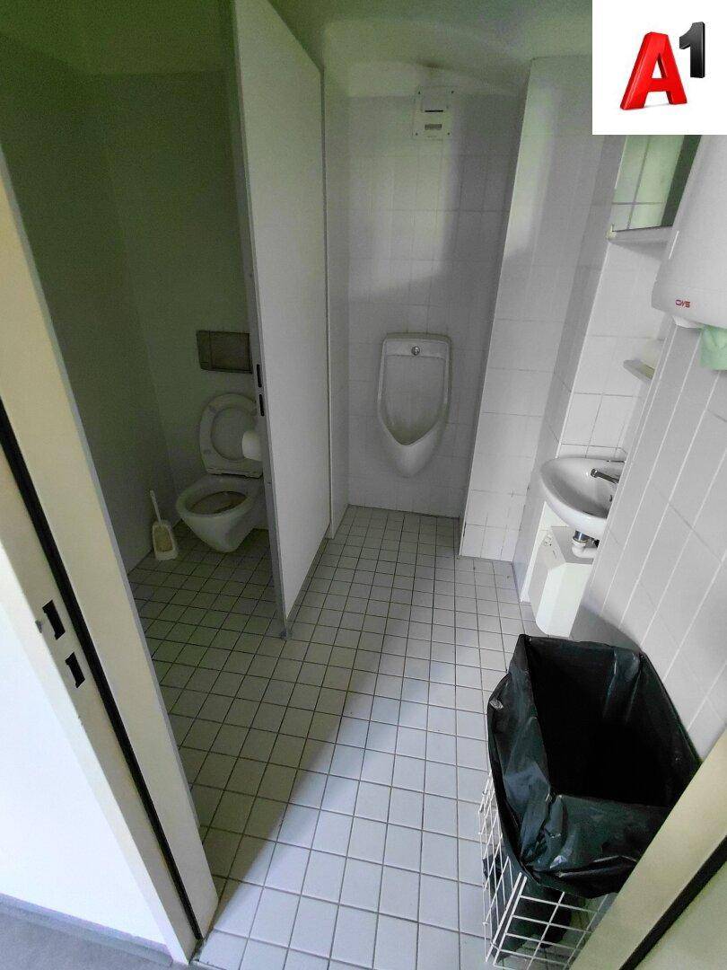 Öffentliche Toilette mit WC-Kabine, Urinal, Waschbecken und weißen Fliesen an Boden und Wänden.