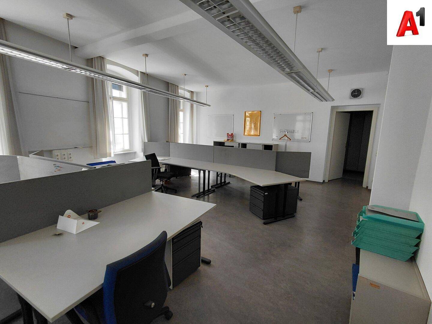 Großraumbüro mit mehreren Arbeitsplätzen, Trennwänden und viel Tageslicht durch die Fenster.
