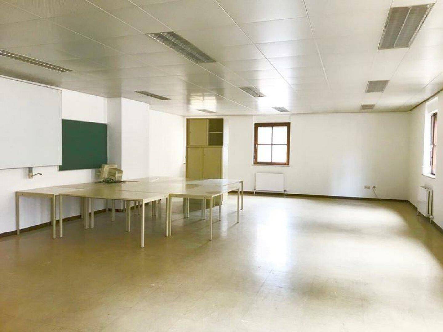 Heller Konferenzraum mit Tischen, einer Tafel und Fenstern, geeignet für Schulungen oder Besprechungen.