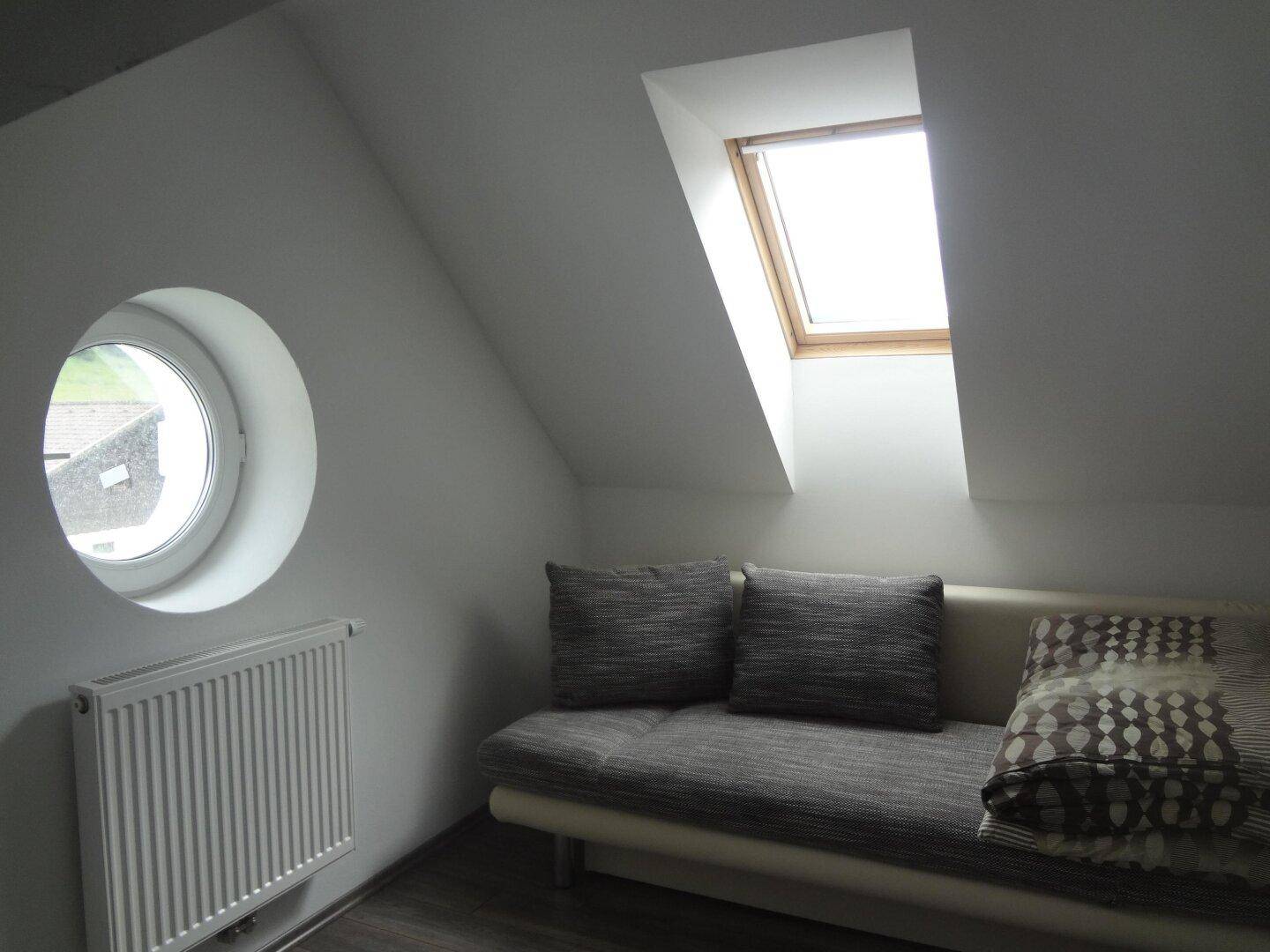 Helles Zimmer im Dachgeschoss mit Schlafsofa, runden und schrägen Fenstern und Laminatboden.