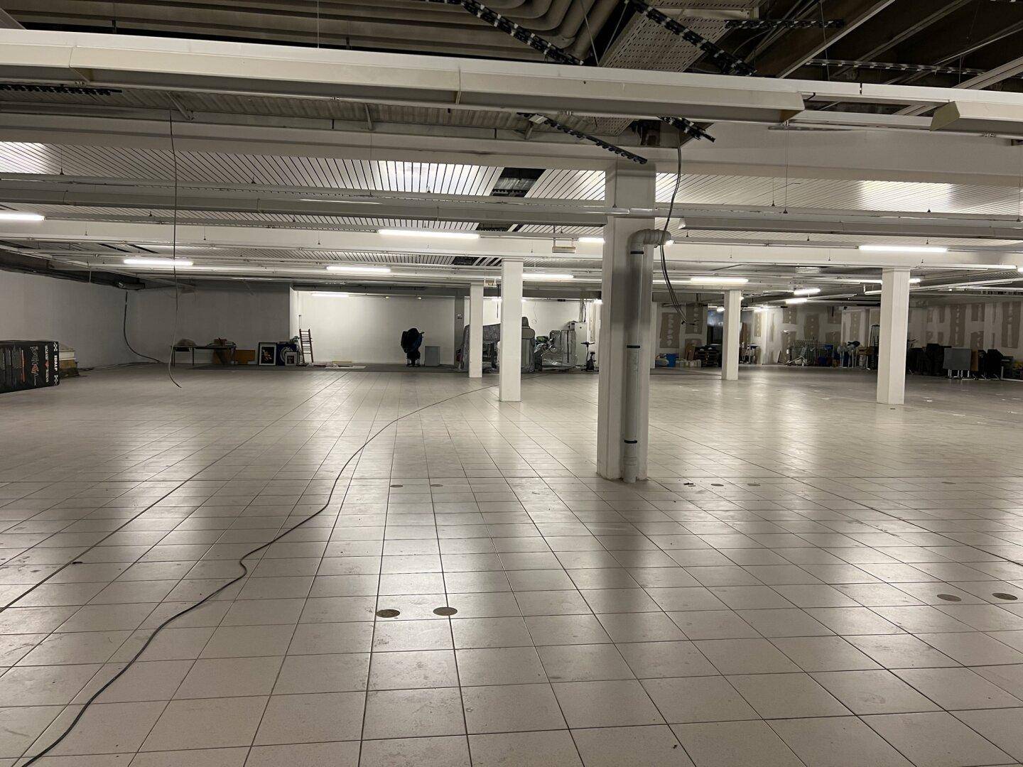 Weitläufiger Gewerberaum mit hellen Fliesen, zahlreichen Stützen und sichtbaren Lüftungsrohren an der Decke.