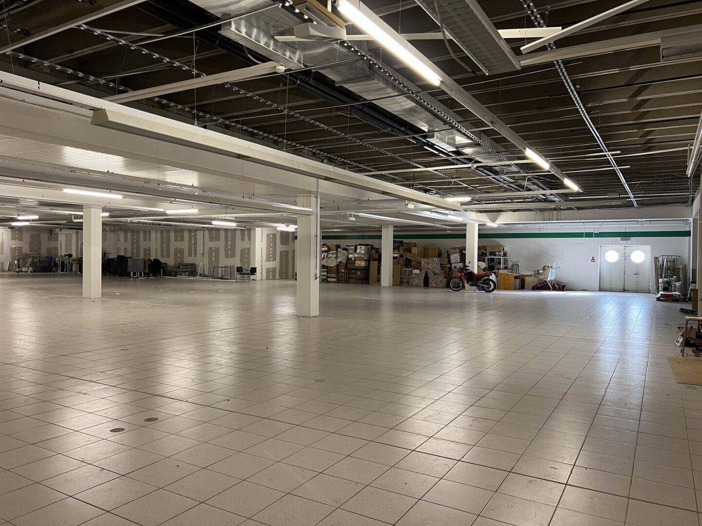 Weitläufiger Gewerberaum mit hellen Fliesen, zahlreichen Stützen und sichtbaren Lüftungsrohren an der Decke.