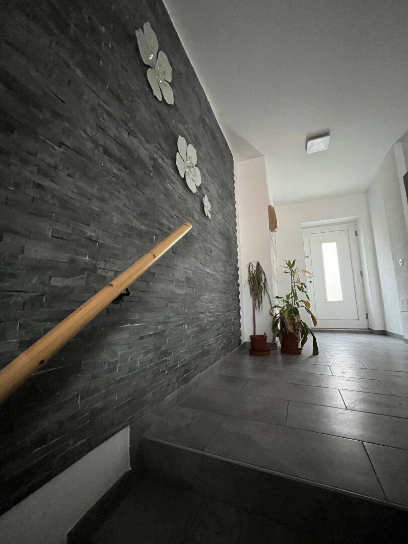 Moderner Eingangsbereich mit dunkler Steinwandverkleidung, Holzhandlauf und grauen Fliesenboden.