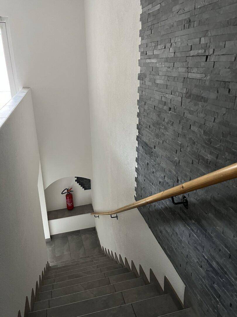 Treppenhaus mit grauen Fliesenstufen, dekorativer Steinwand und Holzhandlauf.