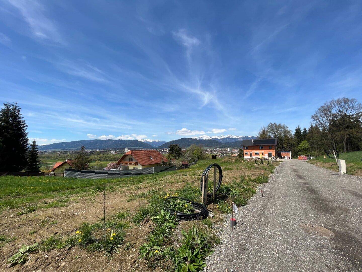 Sonniger Ausblick auf ein Wohngebiet mit Neubauten, umgeben von grünen Hügeln und schneebedeckten Bergen.