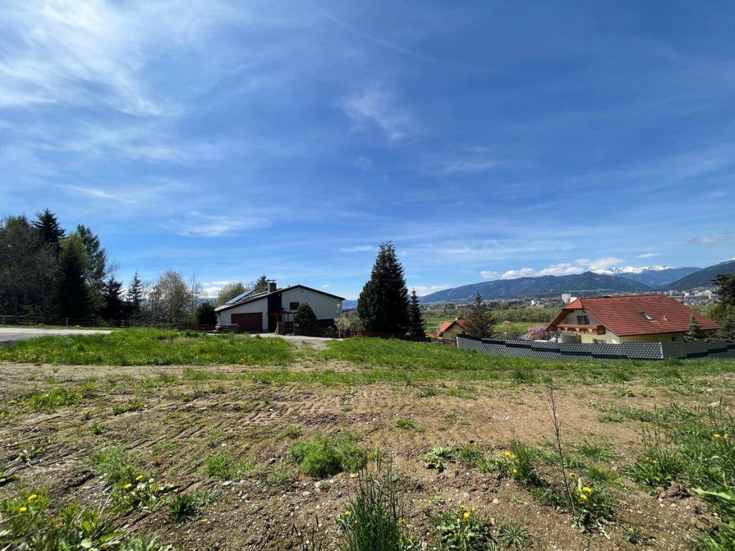 Blick über eine grüne Wiese auf Häuser und die ferne Bergkette unter einem strahlend blauen Himmel.