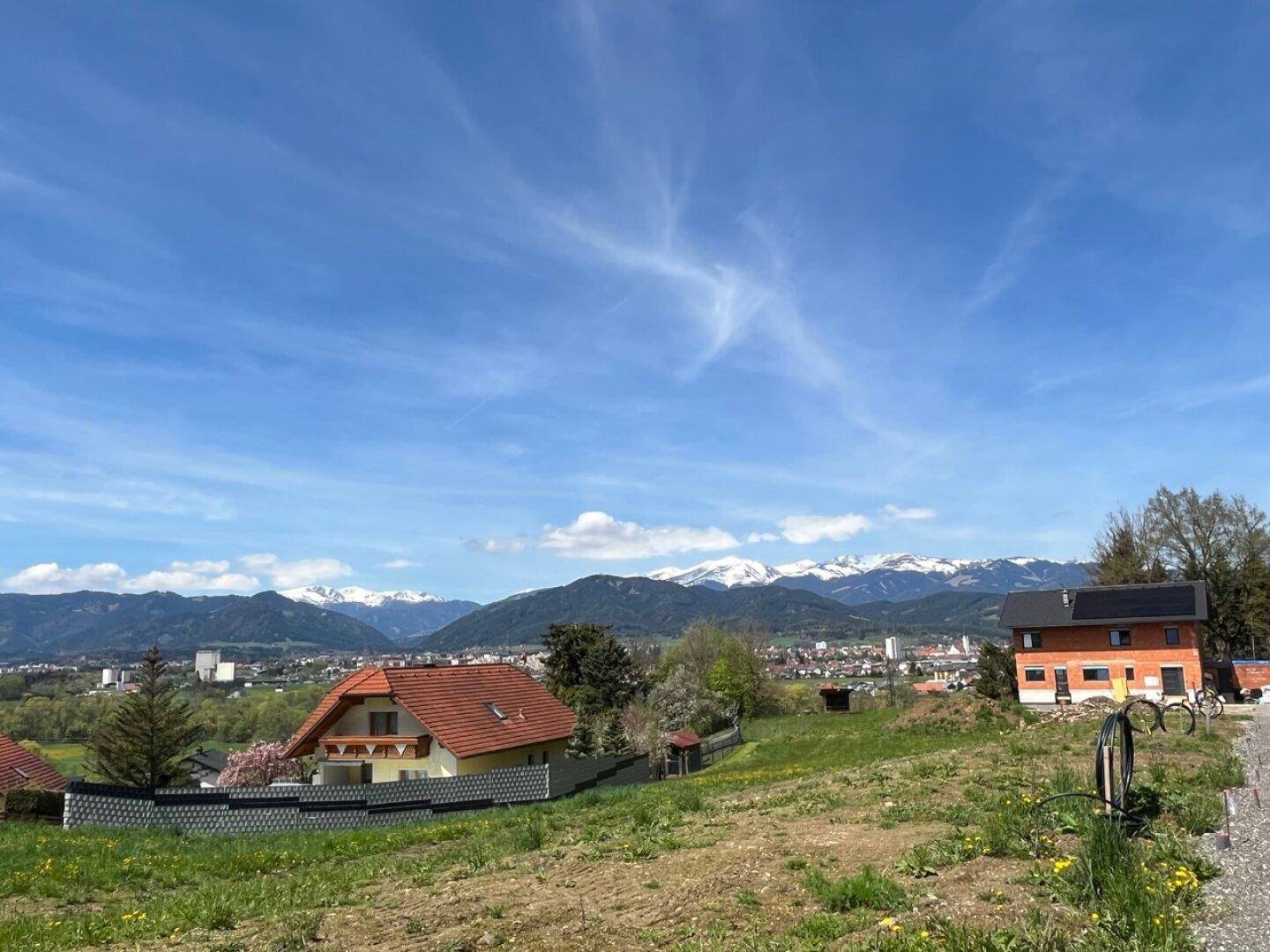 Weitläufiger Ausblick auf die Stadt, grüne Hügel und die majestätischen schneebedeckten Berge am Horizont.