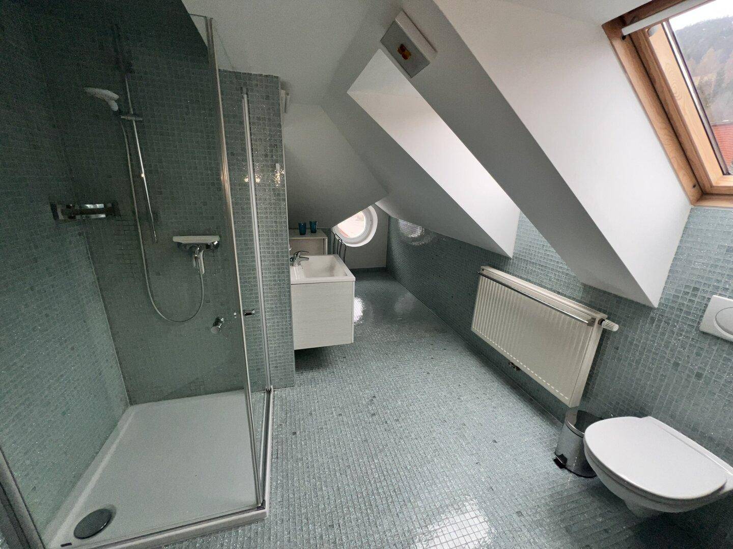 Modernes Badezimmer im Dachgeschoss mit Duschkabine, WC und blauen Mosaikfliesen an Wänden und Boden.