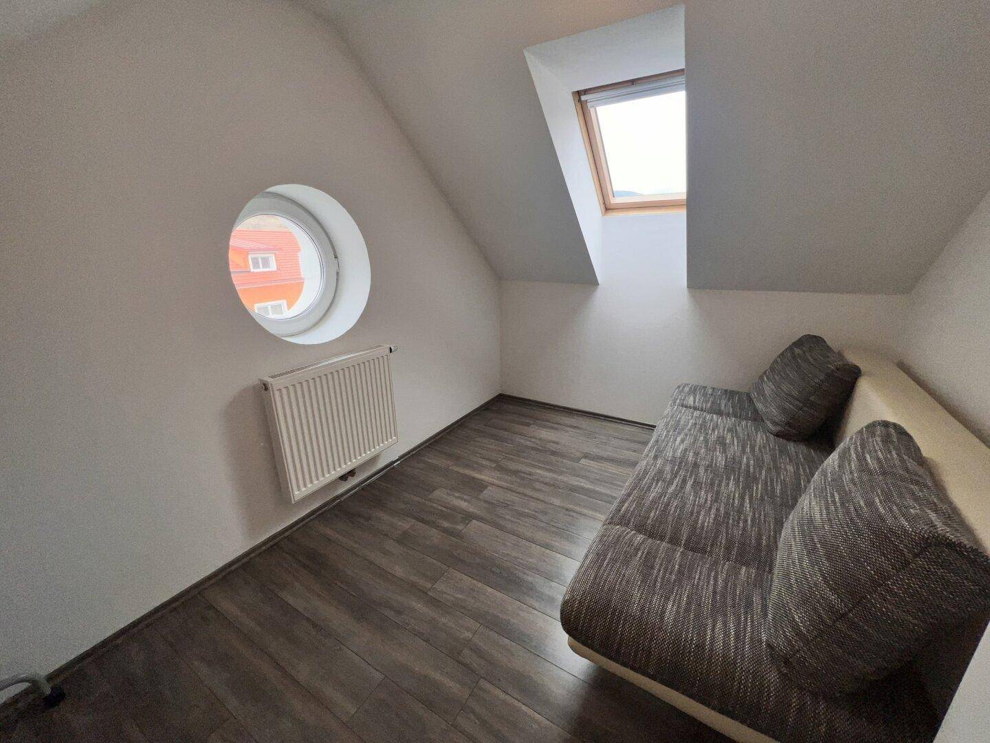Kleines Schlafzimmer im Dachgeschoss mit Schlafsofa, runden und schrägen Fenstern sowie Heizkörper.