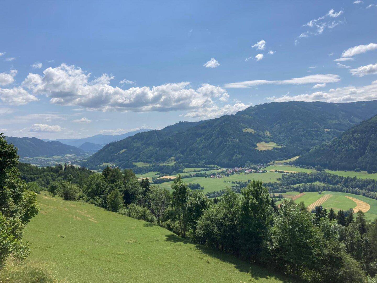 Weitläufige Landschaft mit grünen Hügeln, bewaldeten Bergen und einem Tal unter blauem Himmel mit Wolken.