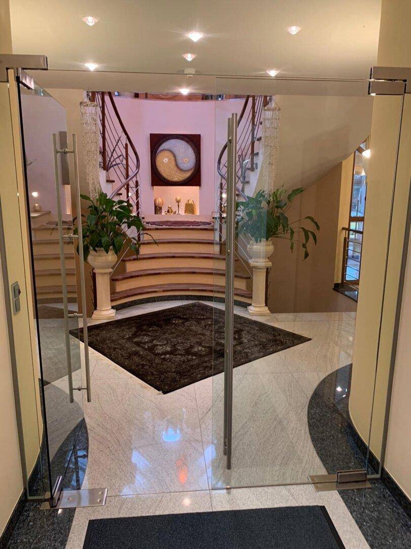 Eleganter Eingangsbereich mit gläsernen Türen, geschwungener Treppe und glänzendem Marmorboden.