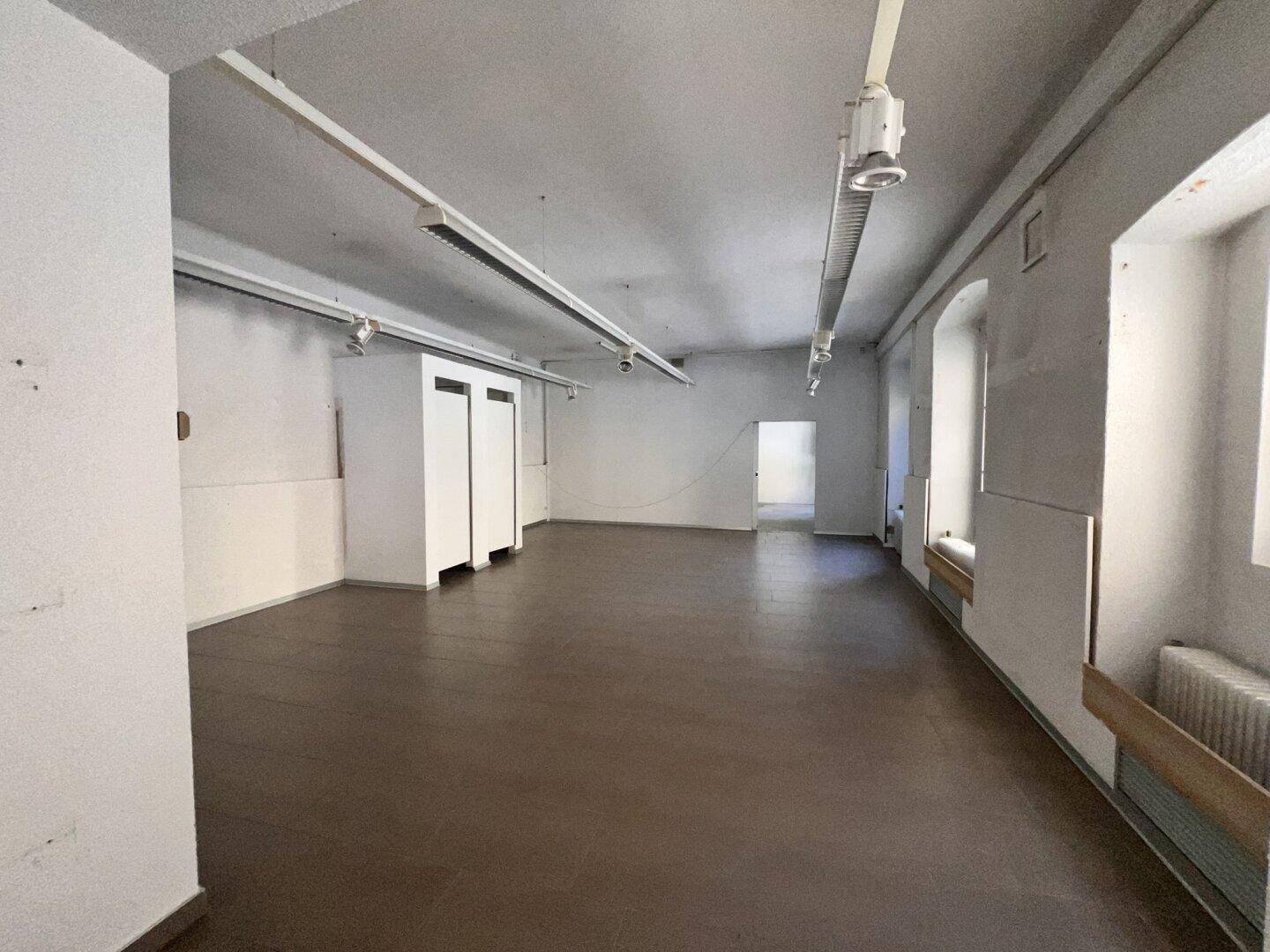 Langer, leerer Gewerberaum mit dunklem Fliesenboden, weißen Wänden und mehreren Fenstern, die Tageslicht spenden.