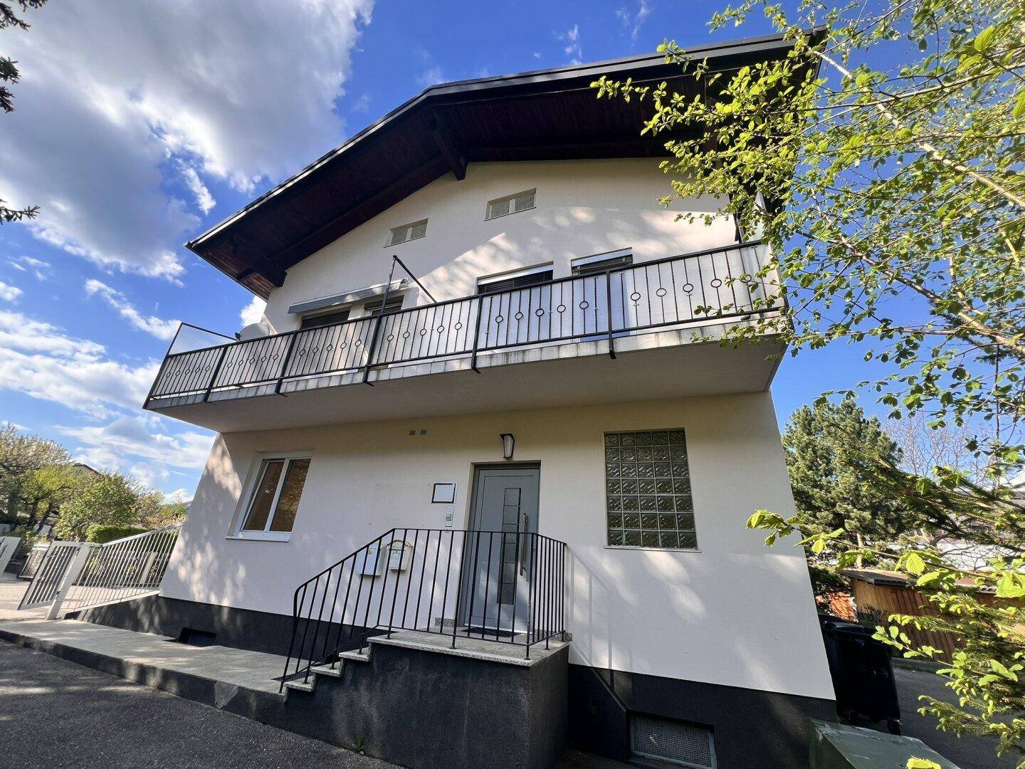 Gebäudeansicht von vorne mit Balkon und Eingangsbereich, umgeben von Bäumen und Grünflächen.