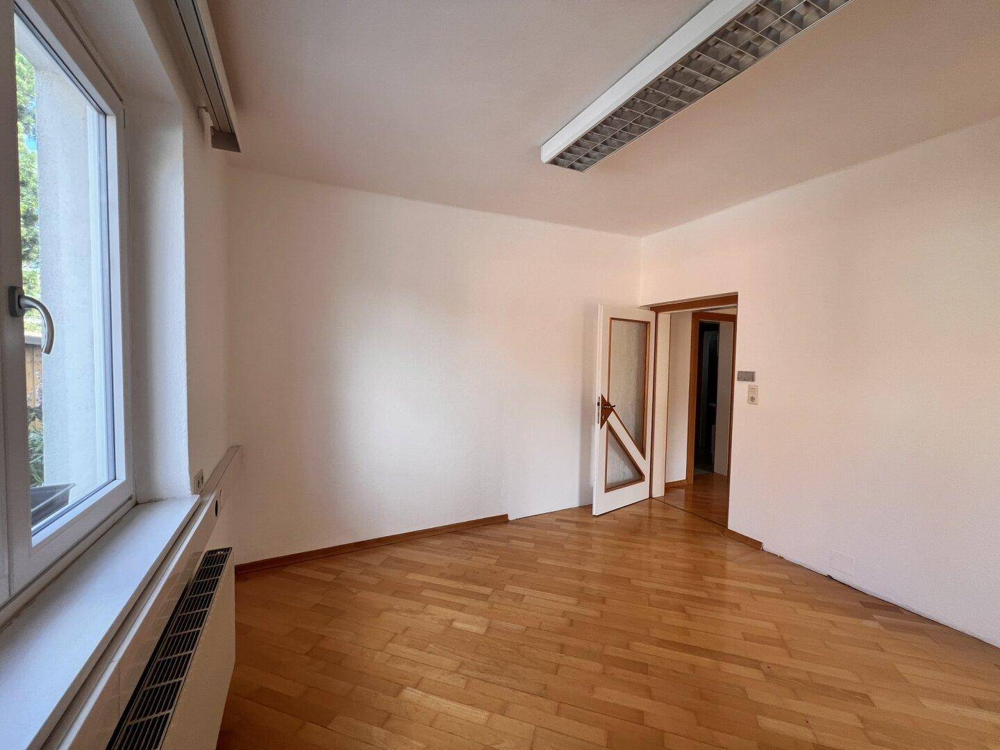 Leerer Büroraum mit Holzboden, einem Fenster und einer Tür, die zu einem weiteren Bereich führt.