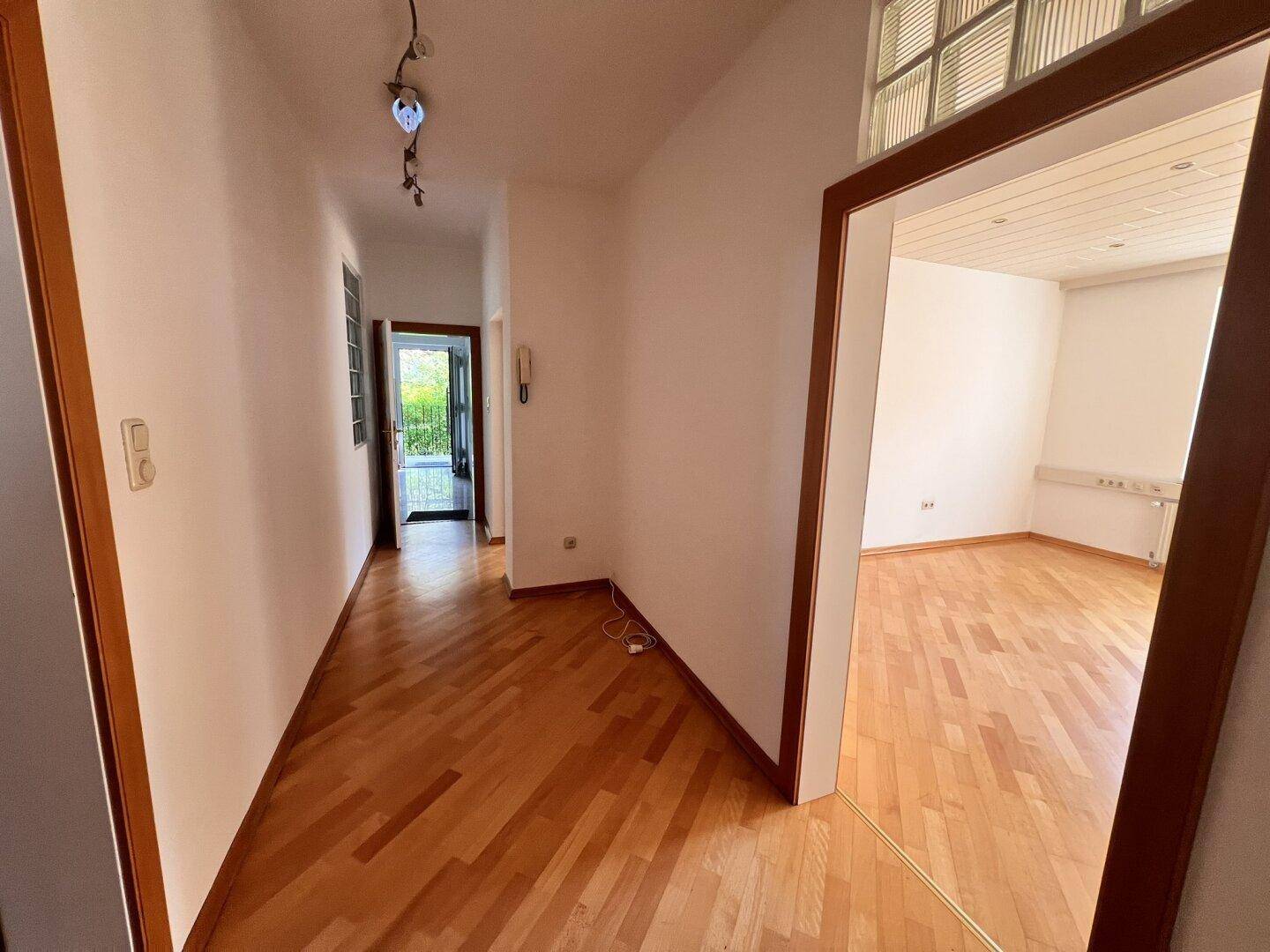 Langer Flur mit Holzboden, der zu einem hellen Raum und einem Ausgang führt.