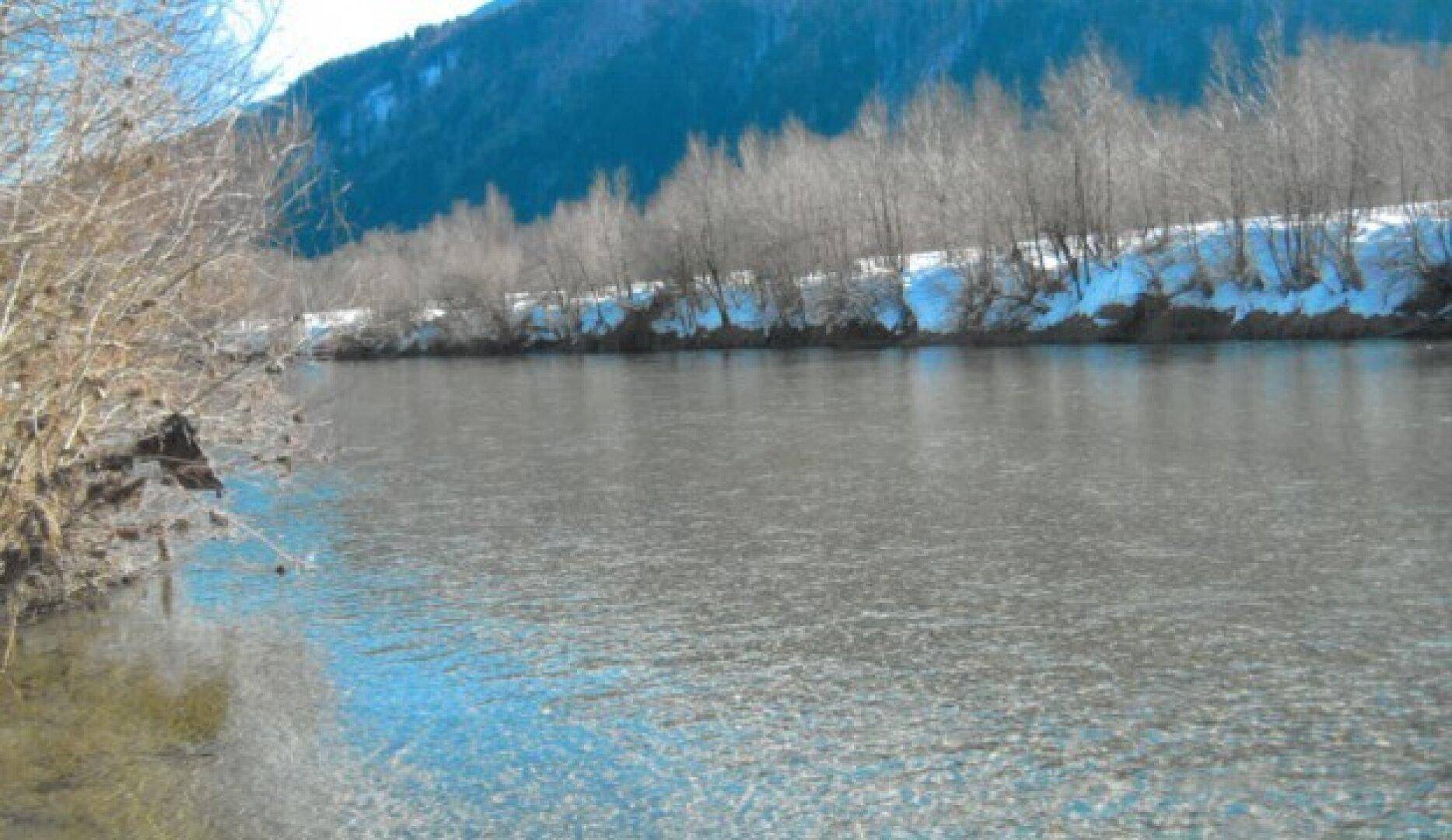 Winterliche Flusslandschaft mit schneebedeckten Ufern und kahlen Bäumen unter blauem Himmel.