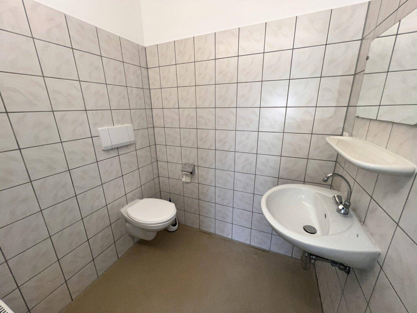 Helles Badezimmer mit weißen Fliesen, einer Toilette und einem praktischen Eckwaschbecken.