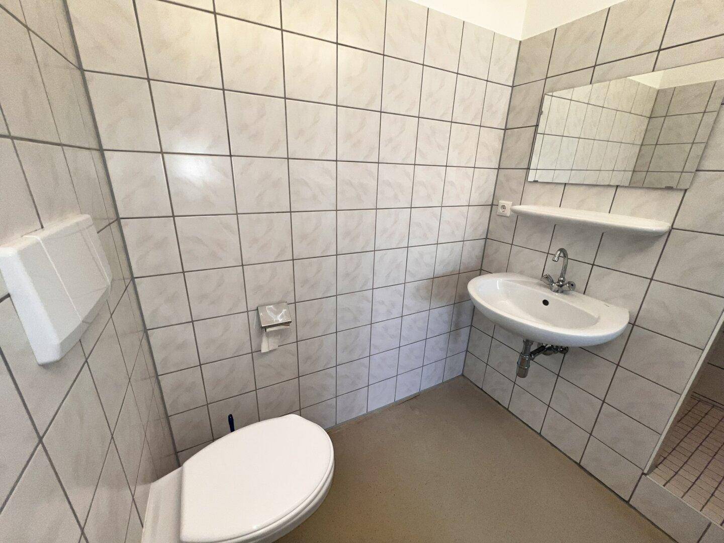 Funktionales Badezimmer mit weißen Fliesen, Toilette, Eckwaschbecken und Duschbereich.