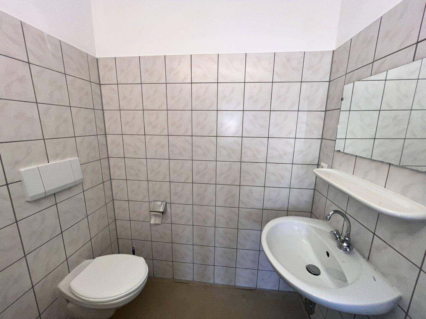 Sauber gefliestes Badezimmer mit Toilette, Eckwaschbecken und einem kleinen Spiegel.