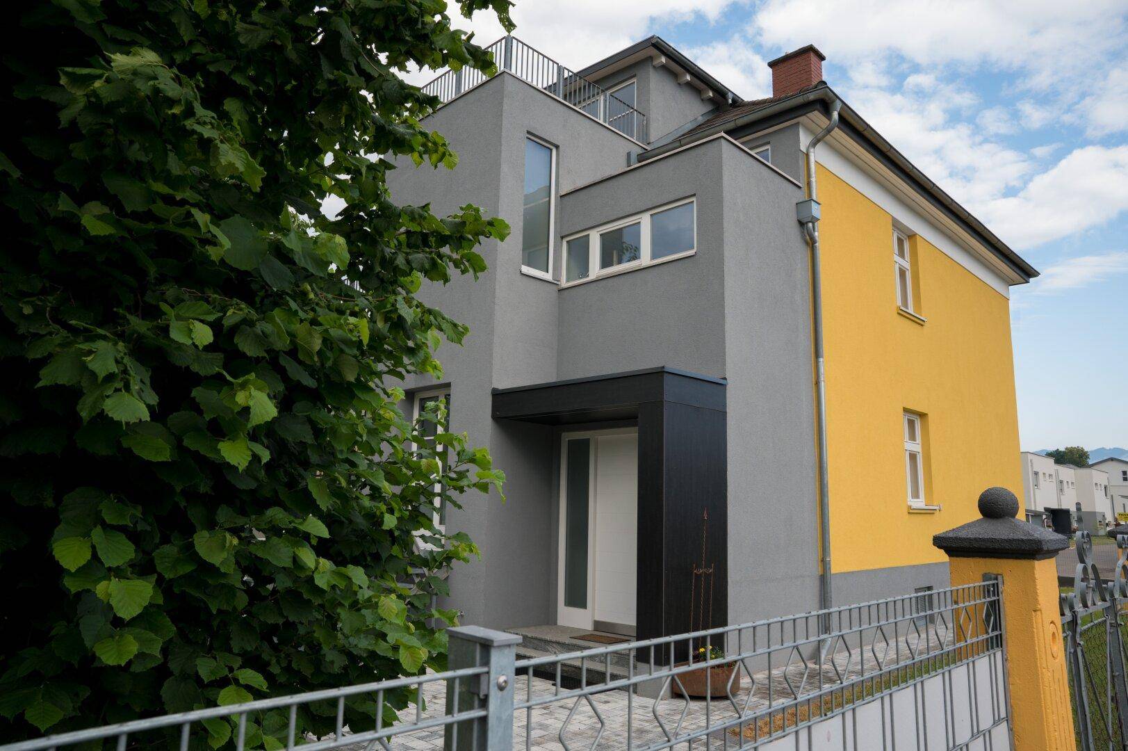 Seitenansicht des Hauses mit gelber Fassade, grauen Akzenten und einem Metallzaun im Vordergrund.