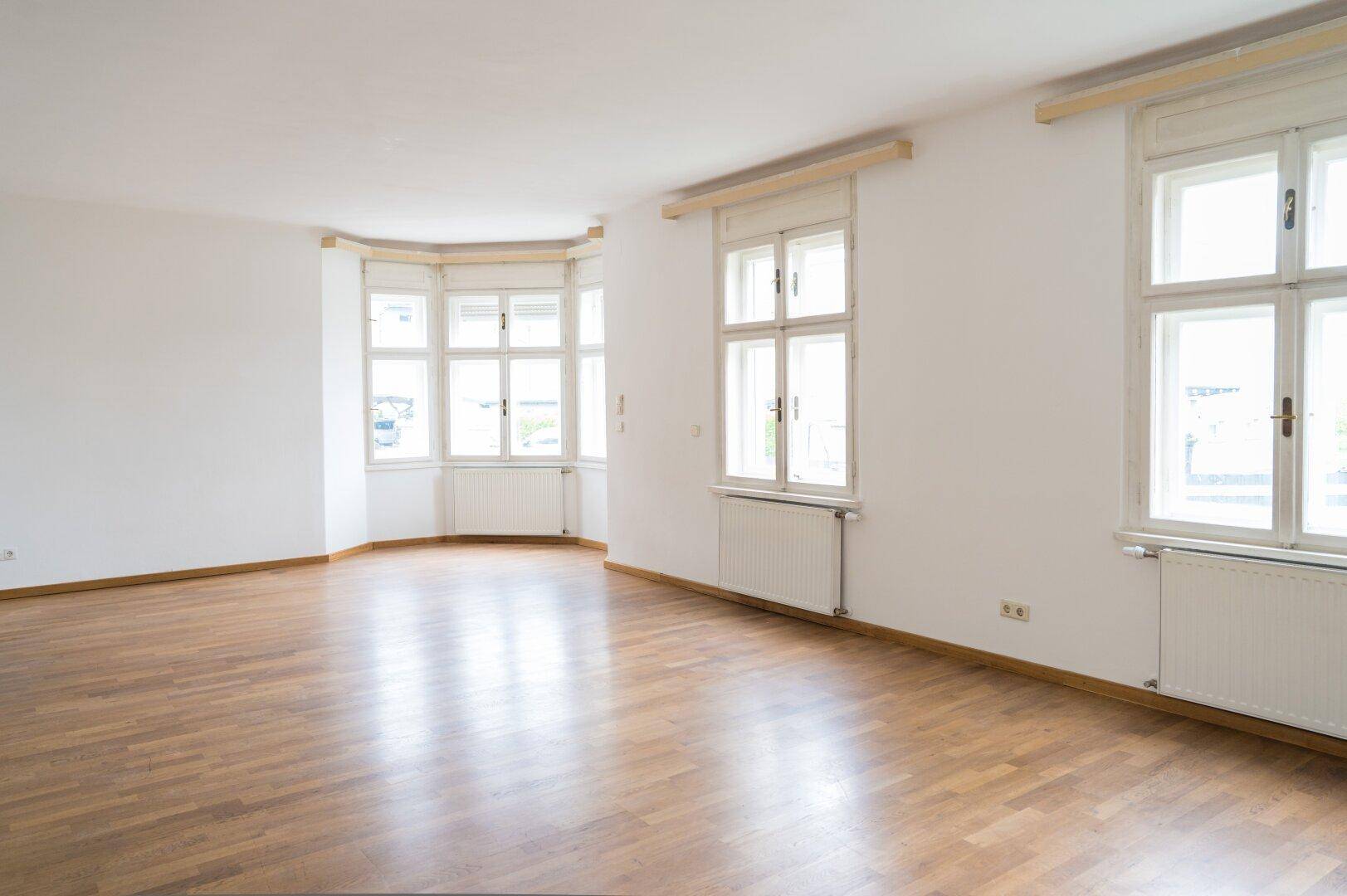 Helles Wohnzimmer mit Holzboden und mehreren Fenstern, die viel Tageslicht hereinlassen.