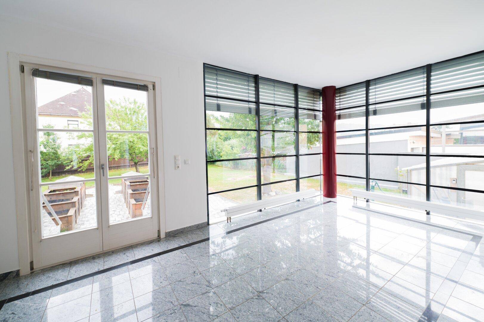 Heller, leerer Raum mit glänzendem Steinboden und großer Glasfront zum Garten.