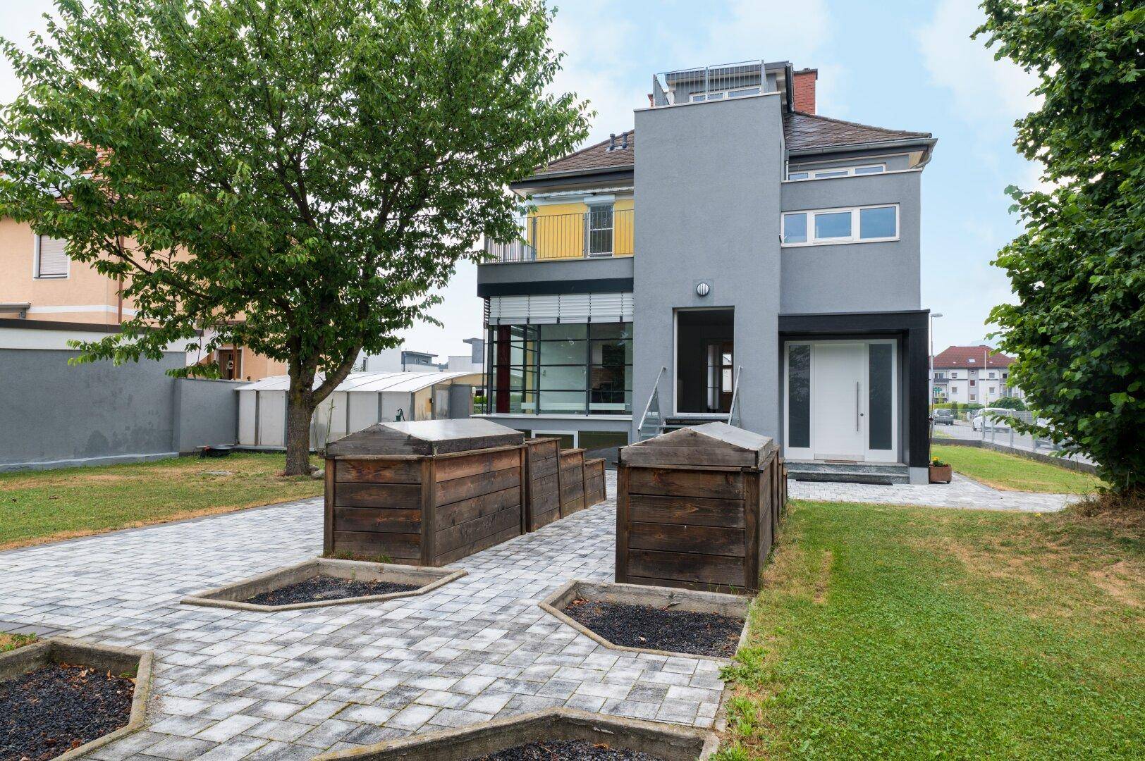 Modernes Wohnhaus mit grauer Fassade, gepflastertem Vorplatz und Hochbeeten im Garten.