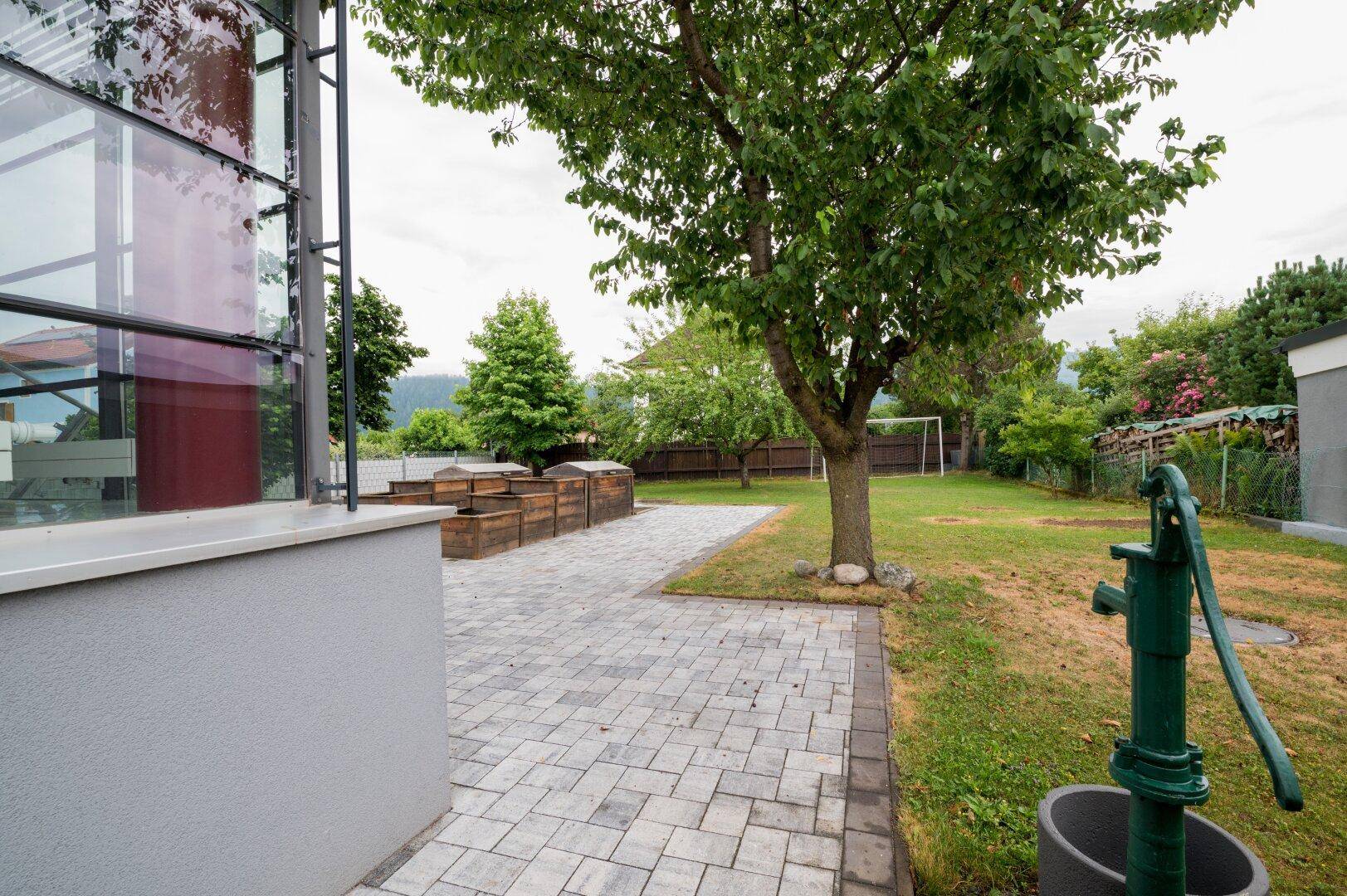 Grüner Gartenbereich mit gepflastertem Weg, alter Wasserpumpe und Blick auf die Berge im Hintergrund.