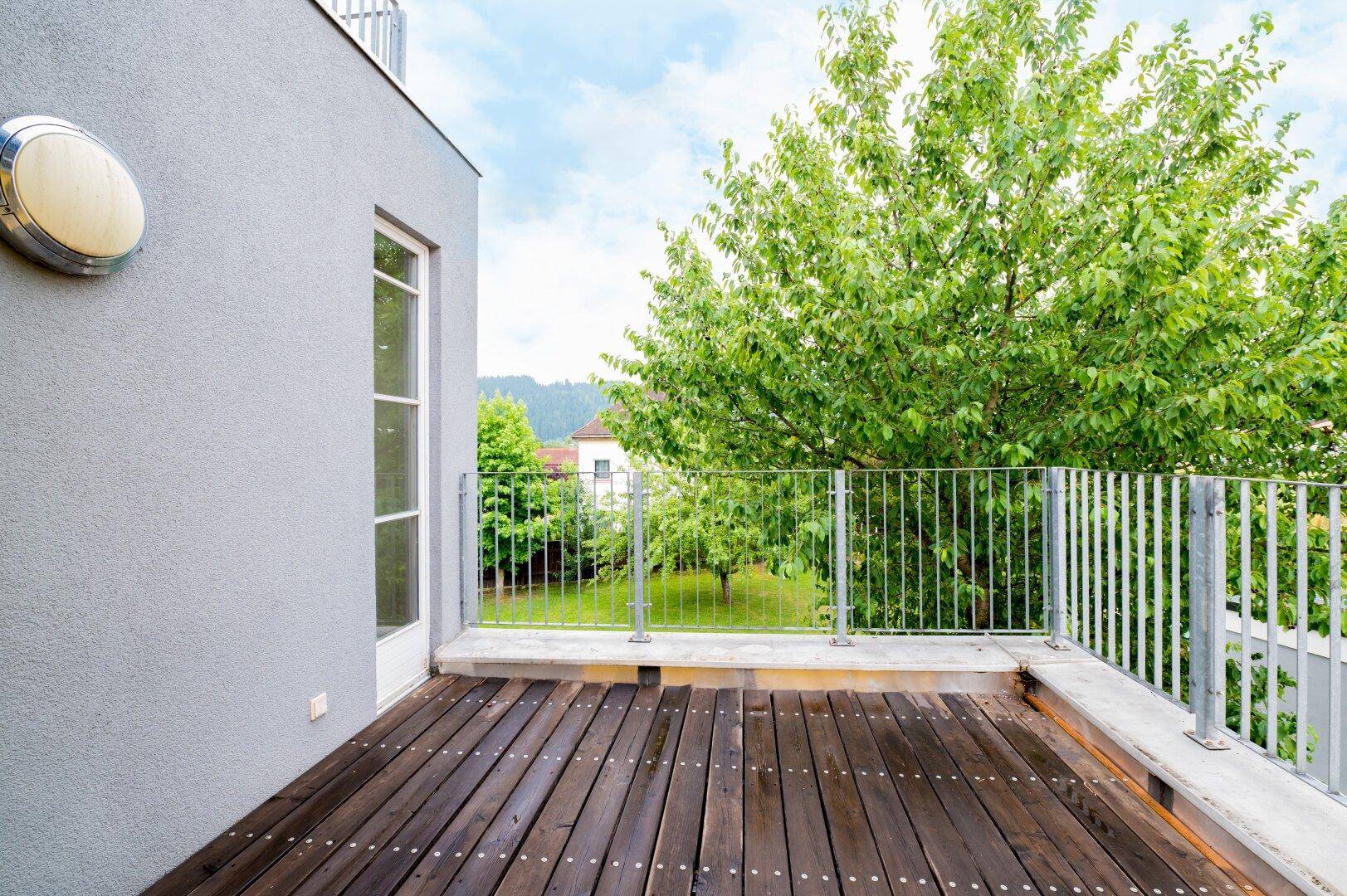 Geräumiger Balkon mit Holzboden und Metallgeländer, umgeben von grünen Bäumen.