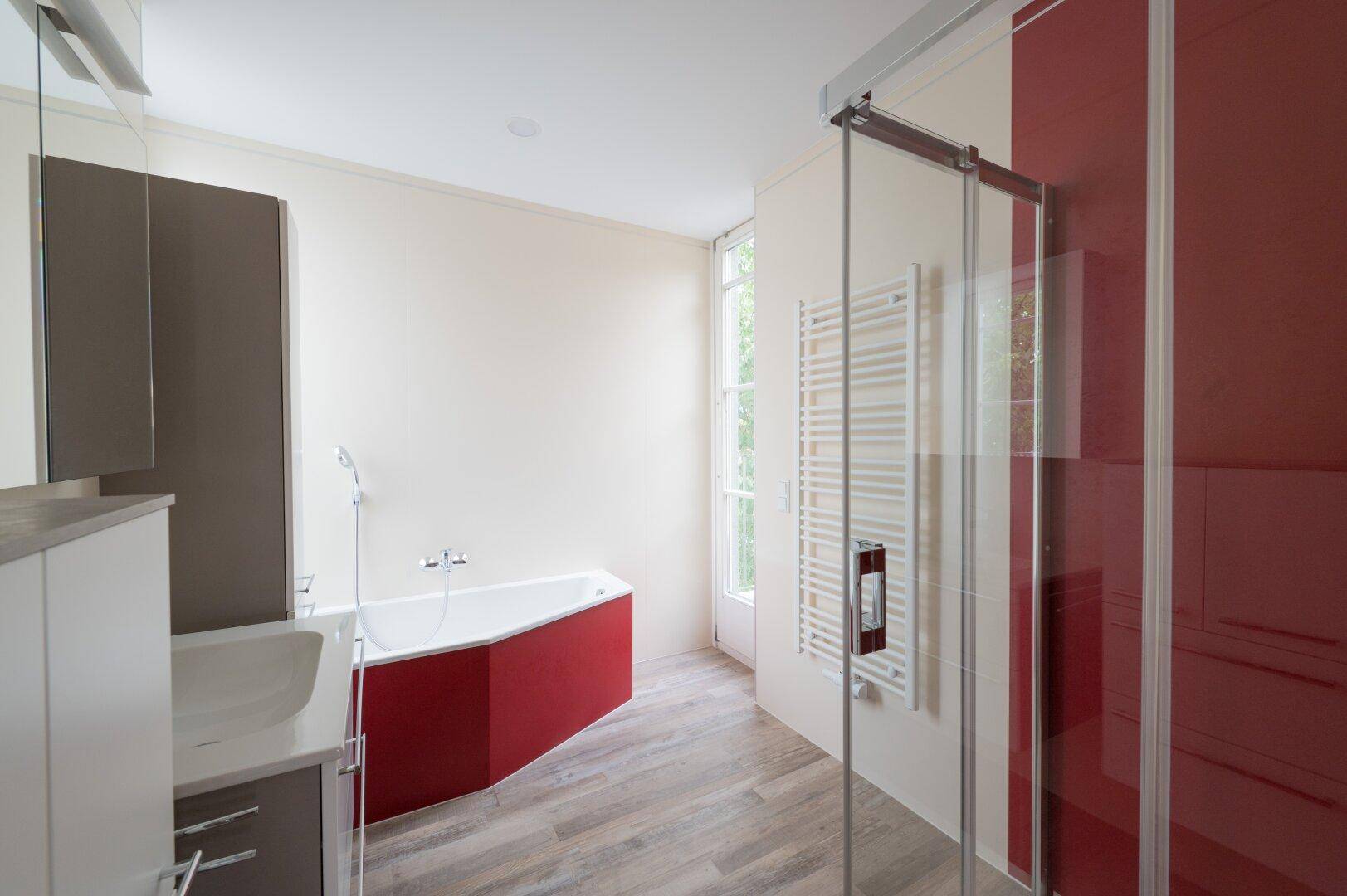 Modernes Badezimmer mit roter Badewanne, Duschkabine und Holzoptik-Fliesenboden.