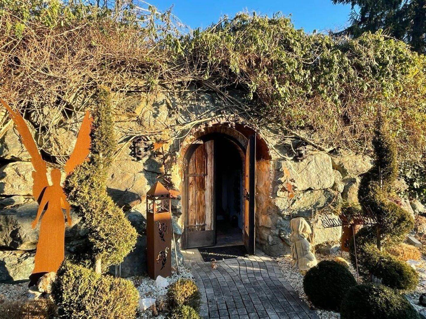 Rustikaler Steineingang zu einem Keller oder einer Grotte, umgeben von üppiger Vegetation.