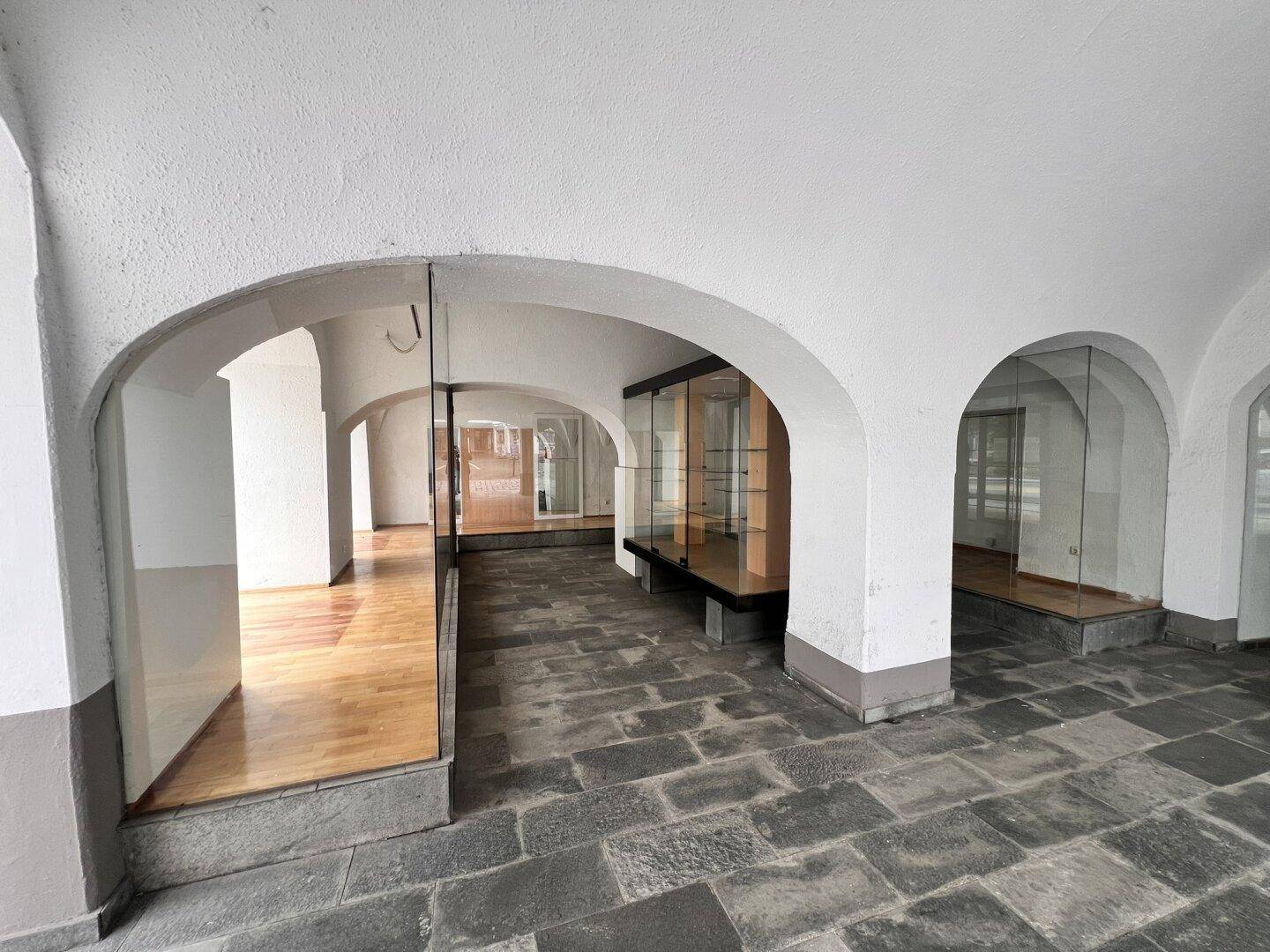 Historische Arkaden mit Steinboden und modernen Glasfronten, die in Ladenflächen führen.