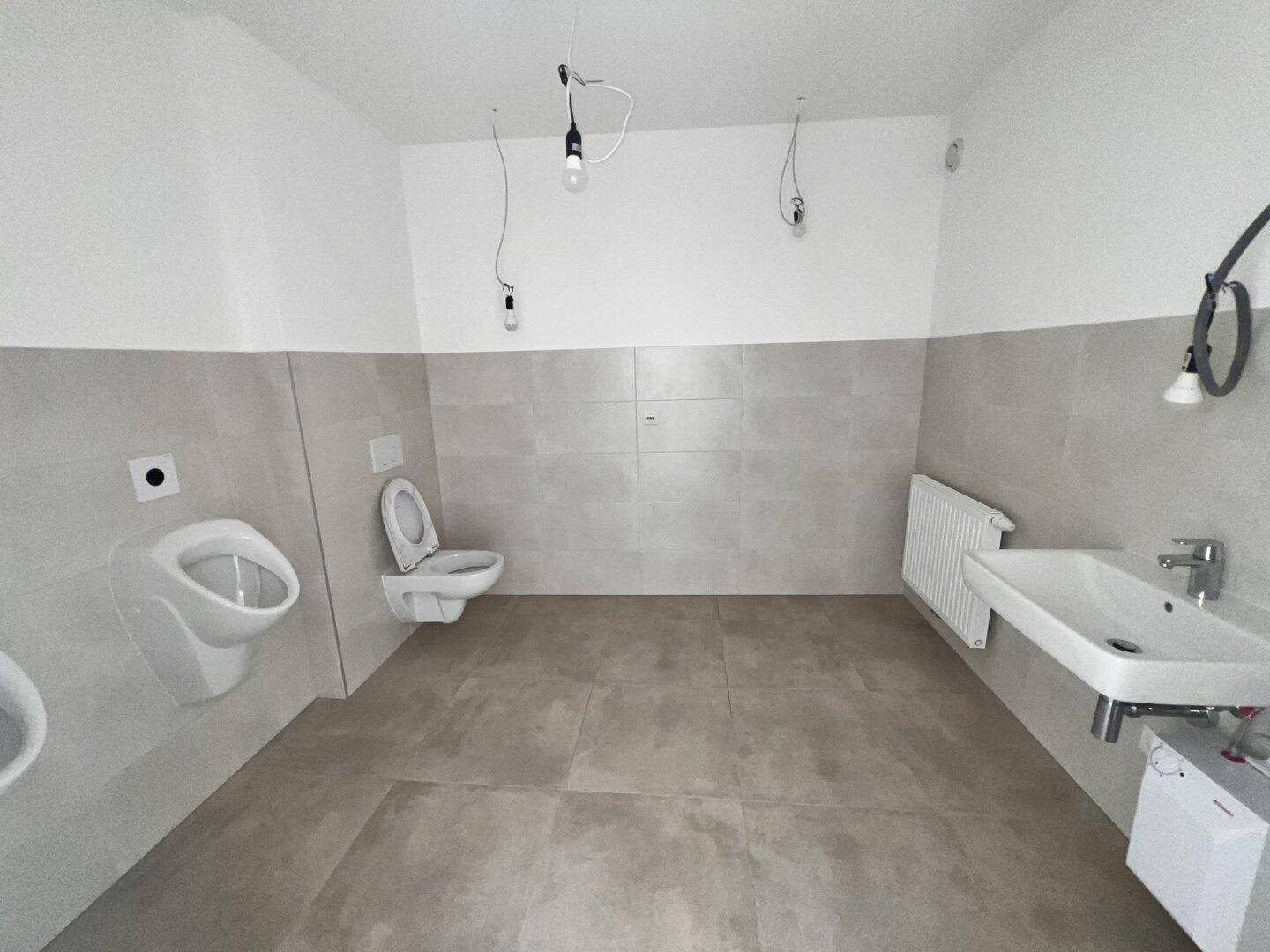 Geräumiges WC mit Urinal, wandhängender Toilette, Waschbecken und modernen grauen Fliesen.
