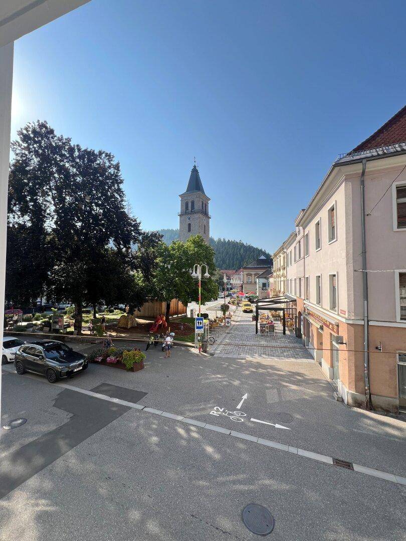 Sonniger Blick auf den belebten Stadtplatz mit Kirchturm, Bäumen und umliegenden Gebäuden.