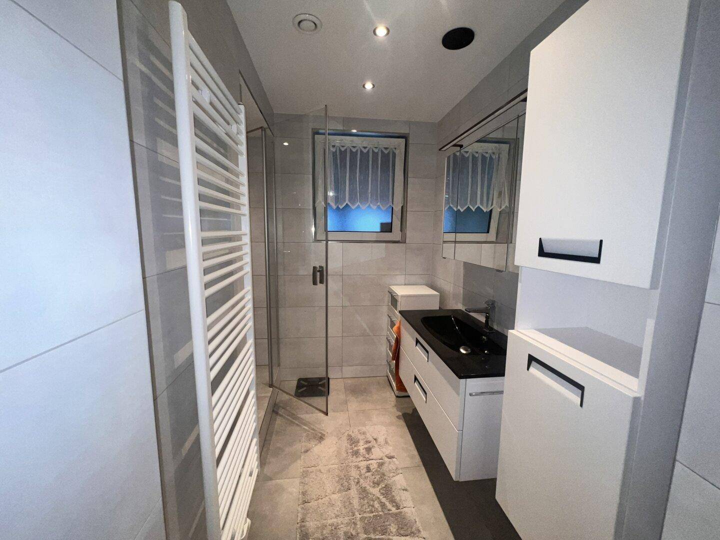 Helles Badezimmer mit moderner Dusche, schwarzem Waschbecken und weißen Schränken.
