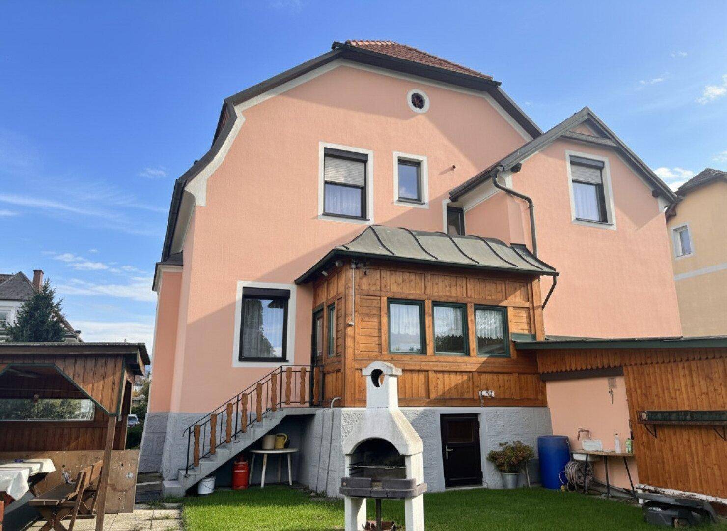 Gartenbereich mit Rasen, gemauertem Grill und Zugang zu einer überdachten Holzterrasse am Haus.