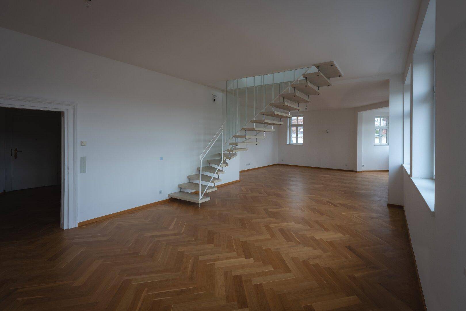 Geräumiges Wohnzimmer mit Fischgrätparkett und einer eleganten Treppe als zentrales Element.
