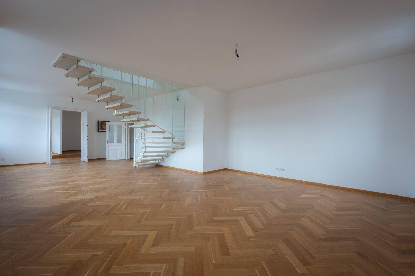 Helles Wohnzimmer mit Fischgrätparkett und einer modernen Treppe, die den Raum strukturiert.