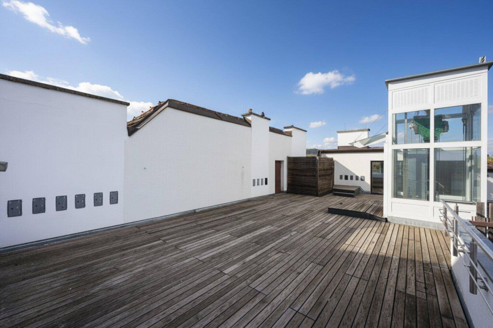 Weitläufige Dachterrasse mit Holzboden und einem gläsernen Aufzug, der den Blick freigibt.