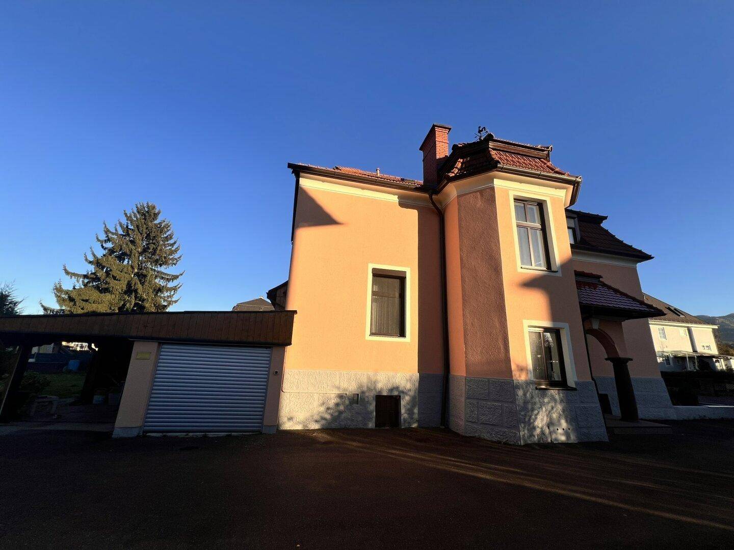 Seitenansicht des Hauses mit rosa Fassade, Garage und Carport unter blauem Himmel.