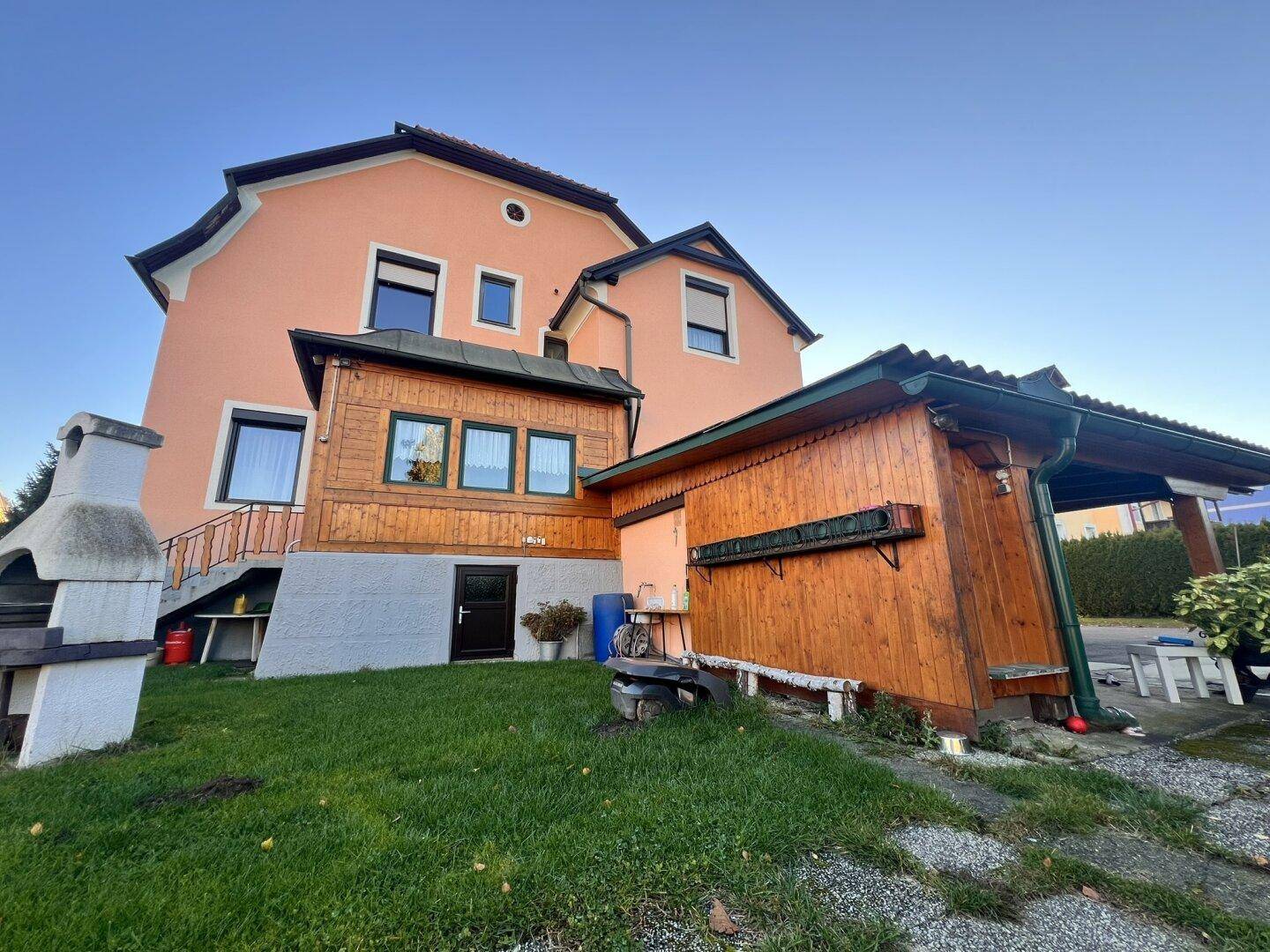 Rückansicht des Hauses mit Garten, Grillplatz und einem Holzschuppen unter klarem Himmel.