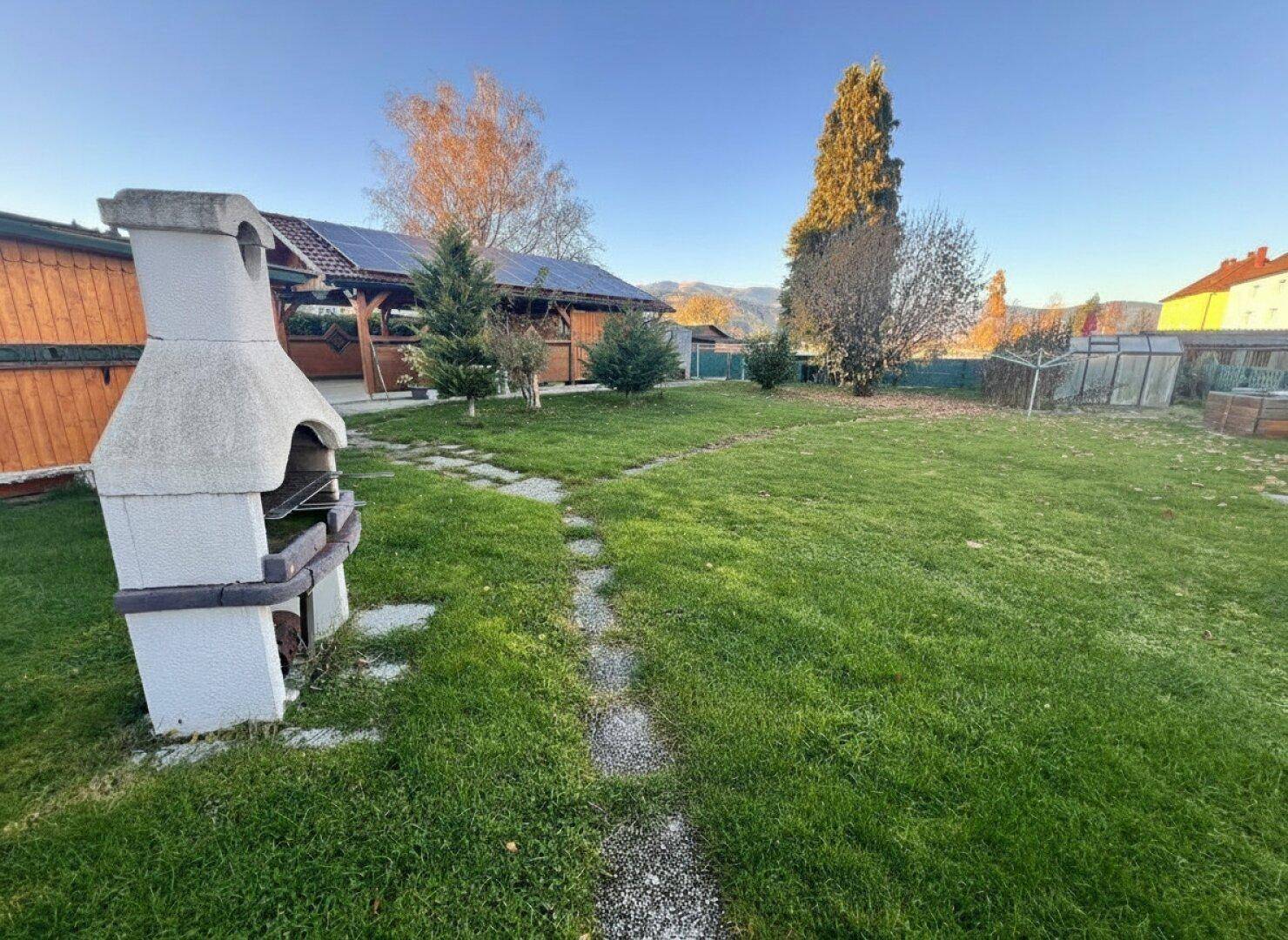 Weitläufiger Garten mit Rasenfläche, Steinweg und einem gemauerten Grill im Vordergrund.