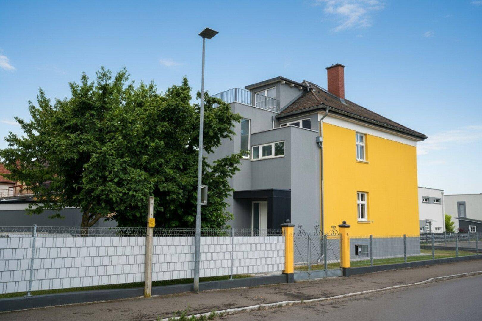 Ansicht des Hauses von der Straße mit gelber und grauer Fassade sowie einem modernen Anbau.