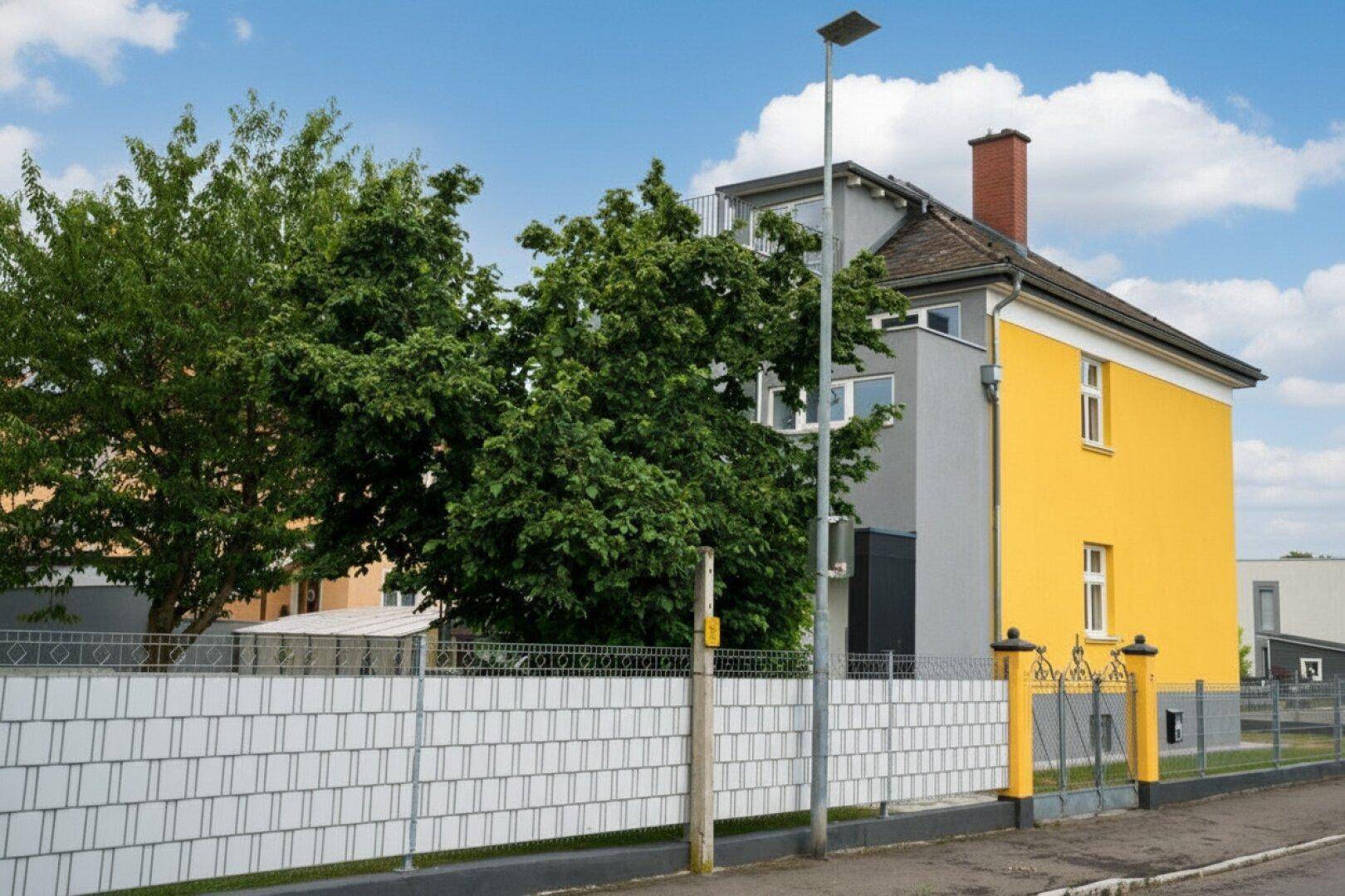 Seitenansicht des Hauses mit gelber und grauer Fassade, umgeben von einem Zaun und Bäumen.