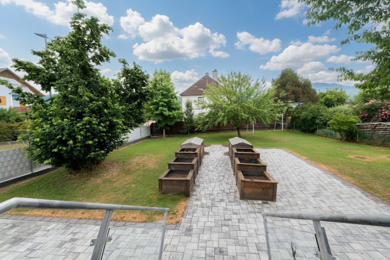 Gepflegter Gartenbereich mit Rasen, Hochbeeten und Bäumen unter blauem Himmel.