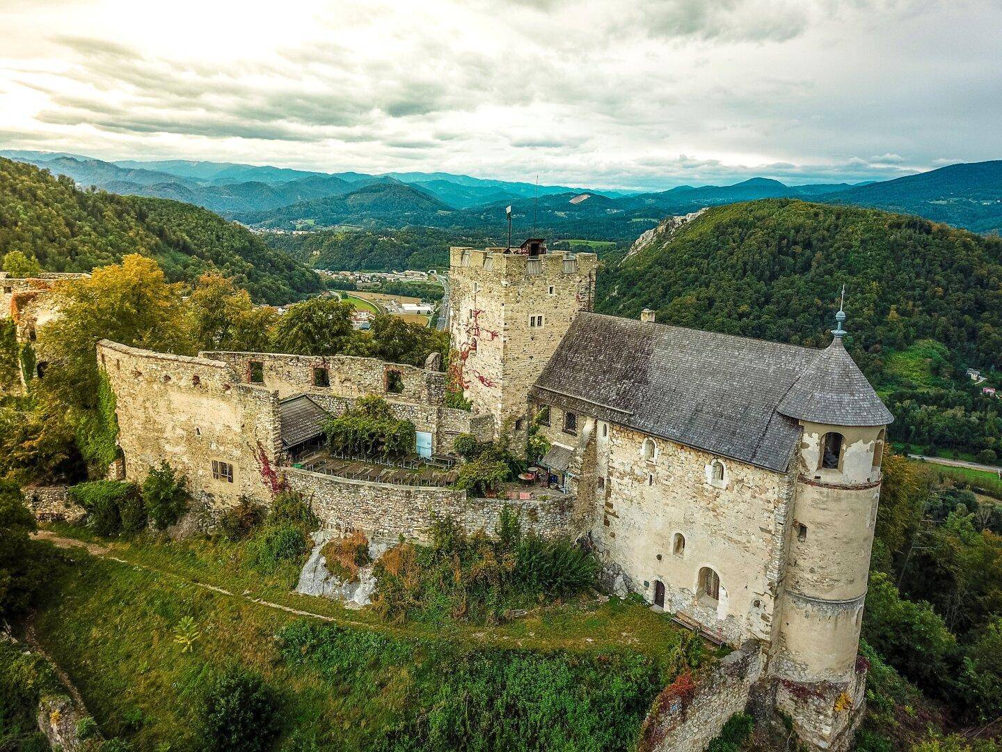 Panoramablick auf eine ausgedehnte Burgruine, eingebettet in eine grüne Hügellandschaft mit Bergen im Hintergrund.
