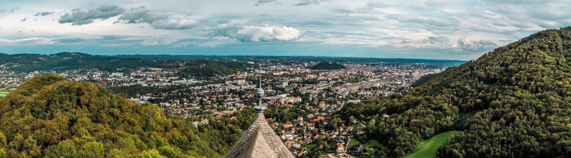 Weitläufiges Panorama einer Stadt, umgeben von bewaldeten Hügeln unter einem bewölkten Himmel.