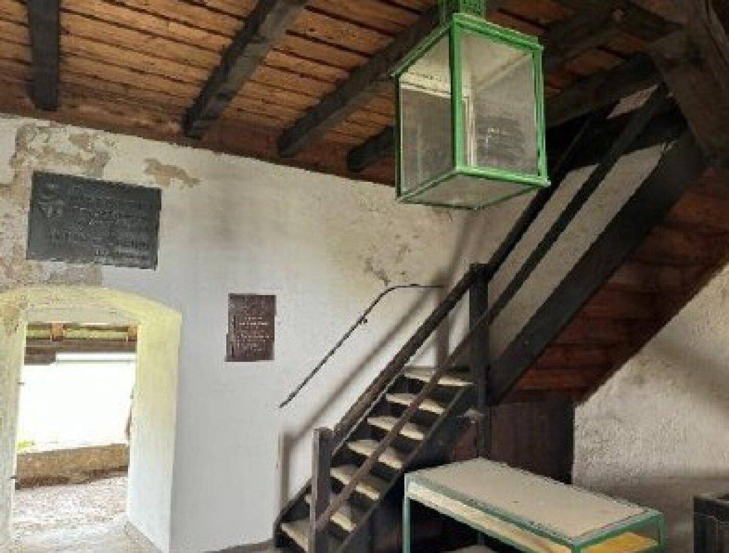 Rustikaler Innenraum einer Burg mit Holzbalkendecke, Steintreppe und einem großen grünen Laternenlicht.